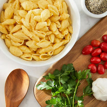 Yellow shell pasta in white bowl on wooden board with tomatoes, parsley, and garlic