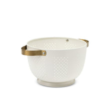 Cream colander with gold strap handle and dotted perforations on bowl.
