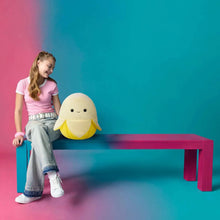 Yellow banana-shaped plush toy with cute face and white belly, sitting on a bench