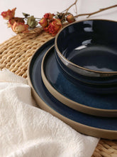 Midnight blue stoneware plates and bowls with gold-trim rims, styled on a woven mat