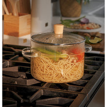 Clear glass pot with metal lid and bamboo knob, handles on both sides, cooking inside.