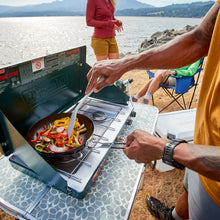 2-burner propane stove with open lid and pan of sautéed vegetables, dark green