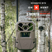 Beige cellular trail camera front view, mounted outdoors with lens window visible.