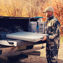 Gray folding table being carried and opened outdoors; black frame and handle visible.