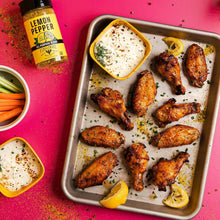Lemon pepper seasoning bottle beside sheet pan of seasoned chicken wings and lemon slices.