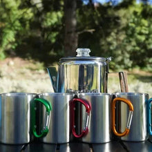 Stainless steel mugs with colored handles and coffee kettle behind them