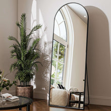 Black arched full-length floor mirror with slim frame, angled beside plants