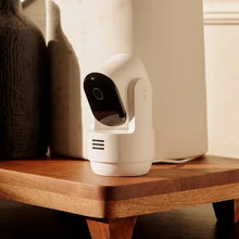 Indoor pan-tilt security camera angled on wooden shelf with white body and black lens