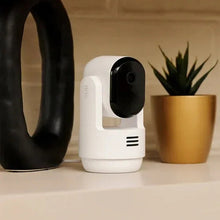 Indoor pan-tilt security camera with white body and black lens, on tabletop among decor
