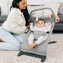 Gray portable baby bouncer with infant secured in seat, caregiver nearby
