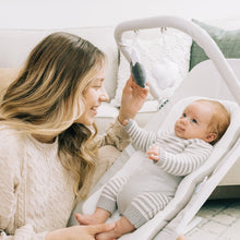 Newborn baby resting in a gray-and-white bouncer while an adult adjusts the seat, side view.