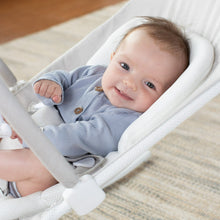 Newborn baby smiling in a light gray-and-white bouncer with patterned cushion.