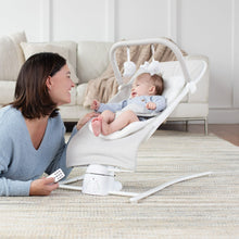 Adult holding a remote beside a newborn in a gray-and-white bouncer on a living room rug.