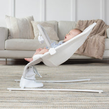 Baby lying reclined in a white-and-gray bouncer with bar supports on a living room rug.