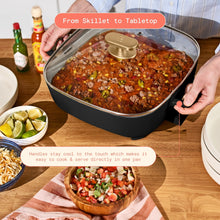 Black electric skillet with glass lid and gold handle, held by its side grips