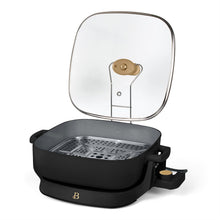 Black electric skillet with glass lid raised and perforated steaming tray inside