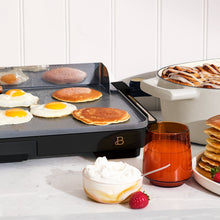XL electric griddle cooking eggs and pancakes, with a black base and gold-tone logo
