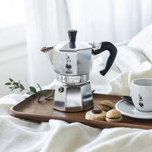 Silver stovetop espresso maker with black handle and knob, displayed on a tray with cup