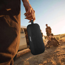 Black portable Bluetooth speaker held in hand outdoors, with carry strap