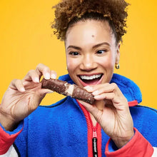 Person smiling while holding and biting a chocolate protein bar