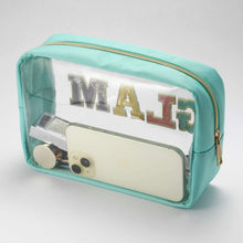Teal cosmetic bag with clear front window, gold zipper, and partial “MA” lettering
