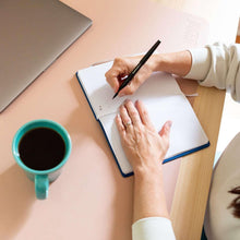 Coffee mug on desk beside notebook and hands writing