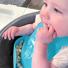 Teal silicone feeding bib in use, baby reaching toward a spoon