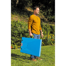 Blue foldable cornhole game board with green handle, carried outdoors
