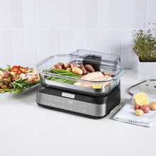 Digital glass steamer with clear lid on countertop, steaming mixed ingredients and greens