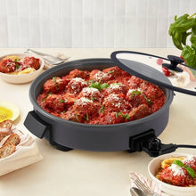 Gray electric skillet with see-through lid, filled with cooked meatballs and herbs