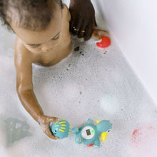 Baby playing with teal dinosaur floating bath thermometer and red toy in bubble bath.