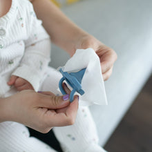 Baby pacifier being wiped clean with white Dr. Brown's wipes held by hands.