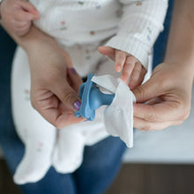 Hands wiping a blue pacifier with a white baby cleaning wipe.
