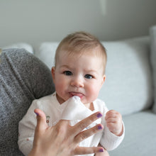 Baby smiling while holding nose and face wipe with caregiver’s hand nearby