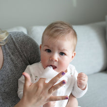 Baby holds a nose and face wipe as caregiver wipes near baby’s mouth