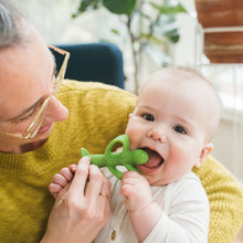 Baby smiling while biting a green silicone peapod teether held by a parent.