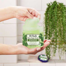 Green shea sugar body scrub jar with hands holding pale green scrub scoop