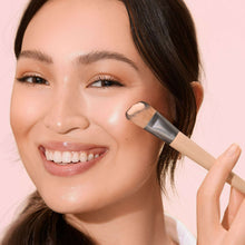 Makeup brush applying cream product near cheek and eye area on a smiling person