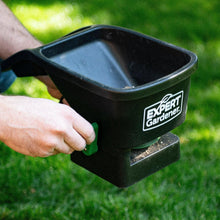 Handheld broadcast spreader held over grass, green lever visible
