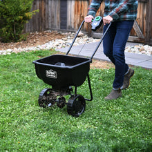 Black push broadcast spreader on lawn with hopper and wheels, operated by person