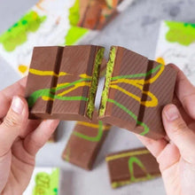 Hands holding two milk chocolate pistachio bars with green filling between layers.