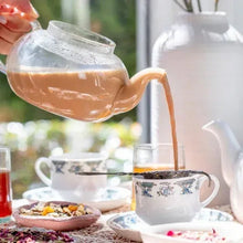 Pouring chai from a glass kettle into a floral teacup with loose tea nearby