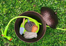 Copper hammered hose storage pot with open lid holding garden tools and hose