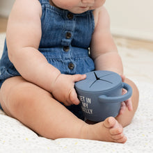Baby sitting holding a light blue snack cup with lid on a cushioned surface.