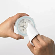 Glass baby food container with light blue seal held open by hands with wiping cloth