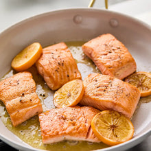 Cream frying skillet with salmon fillets and lemon slices; gold-tone handles