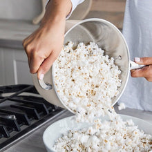 Stainless steel popcorn popper tipped on a stove, pouring popped corn