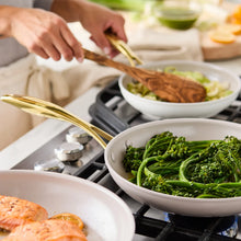 Cream skillet set on gas stove with greens and food in the pans; gold handles