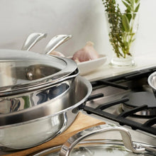 Stainless steel tri-ply cookware pots on gas burners with a wooden board nearby
