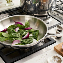 Stainless steel tri-ply sauté pan with handles, cooking greens on gas burner